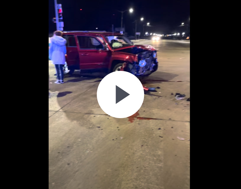 A red SUV with a heavily damaged front end is stopped in an intersection at night. Debris, a small pool of liquid, and some outdoor gear—including a hunting dog kennel—are on the road. A person stands nearby under the traffic lights.