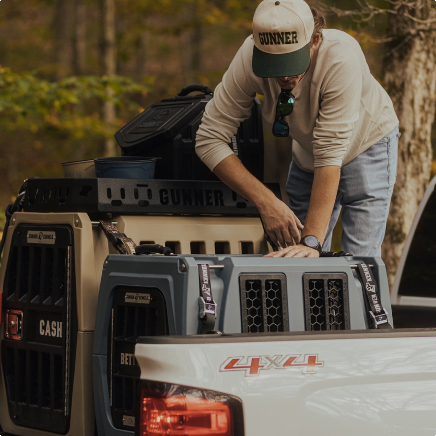 A person wearing a GUNNER cap loads outdoor gear into a 4x4 pickup, next to two large dog kennels labeled CASH and BETTY, in a wooded outdoor setting.