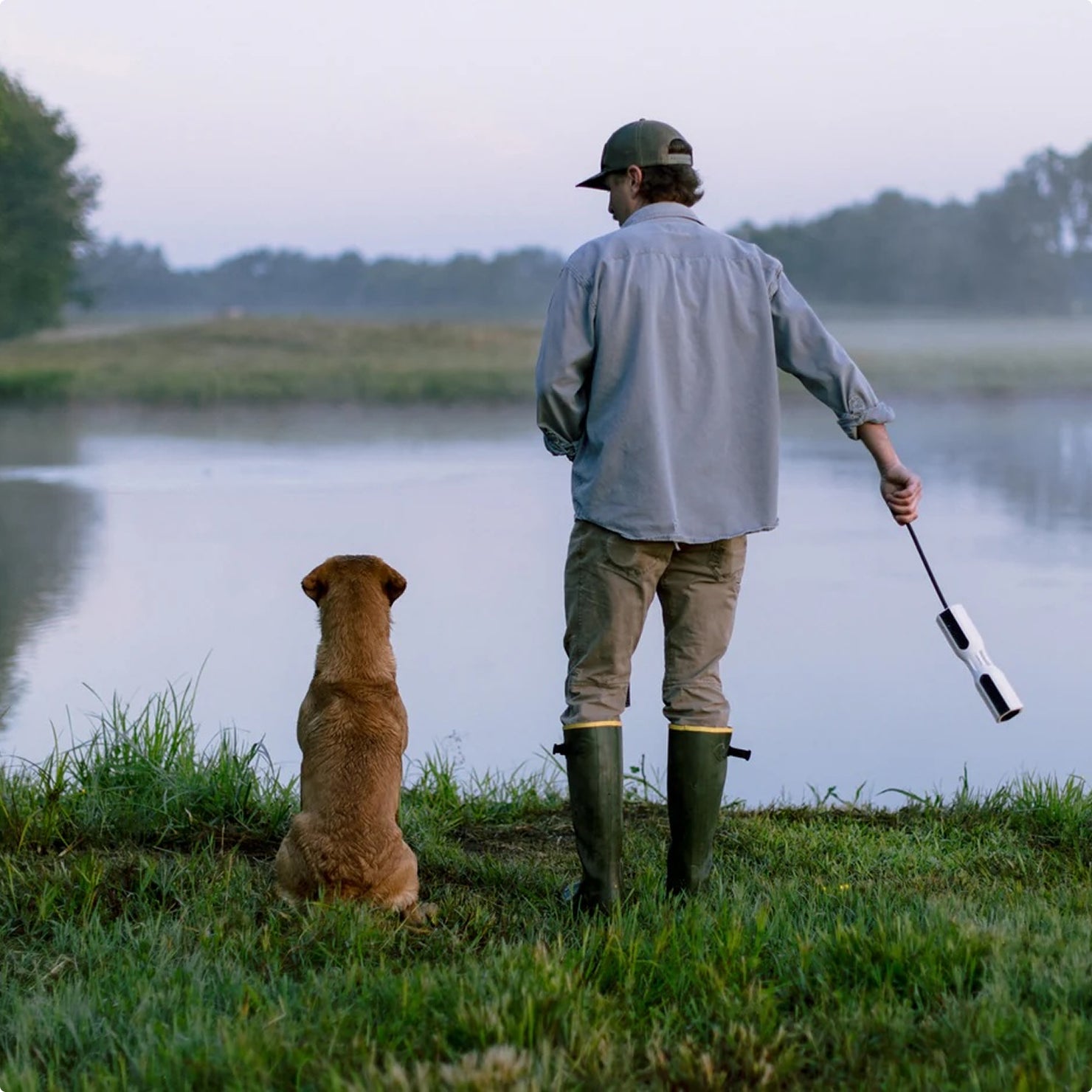 A person in boots and a cap stands by a lake holding a dog training dummy, with a brown dog from the nearby kennel sitting beside them, both facing the water on a misty, calm morning.