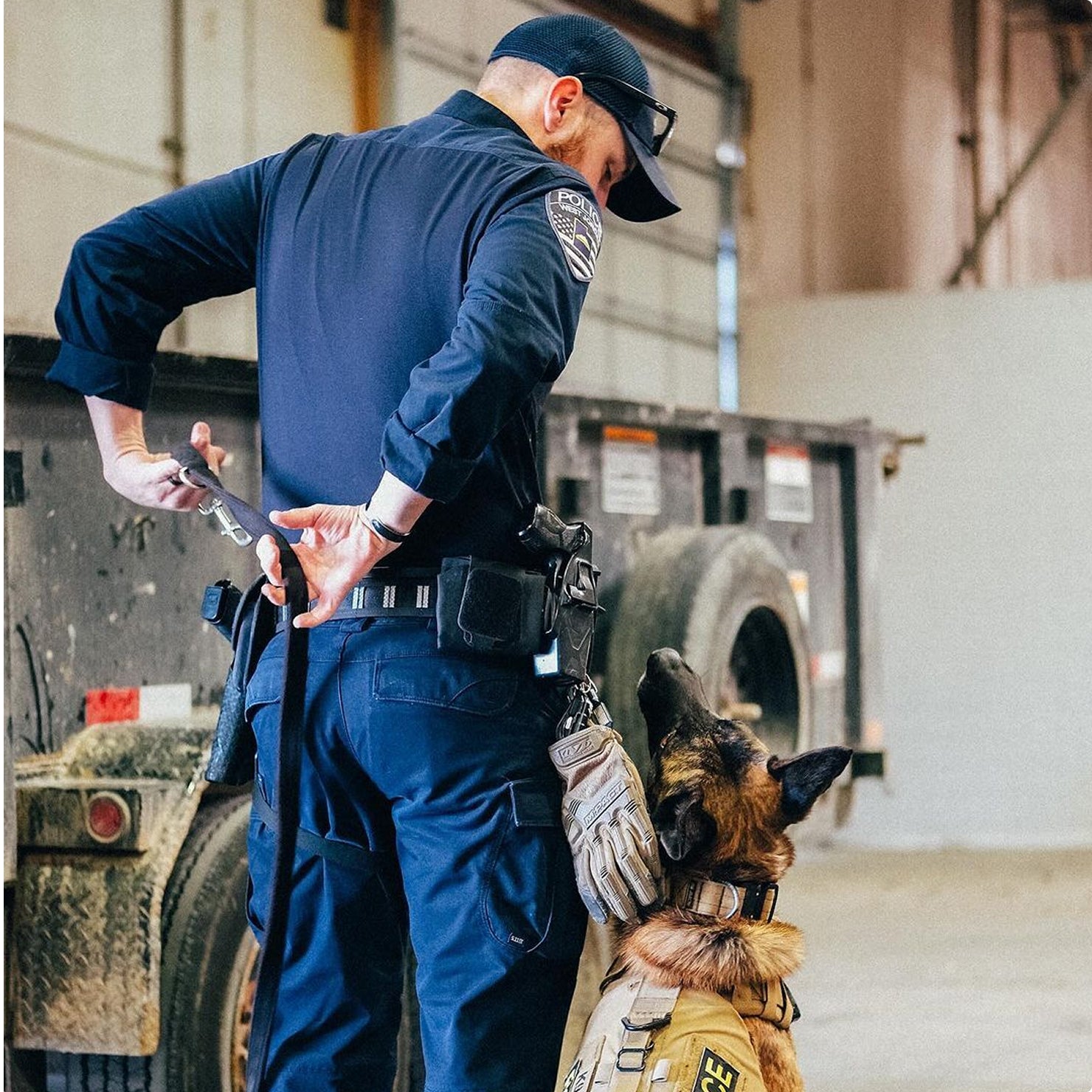 A police officer in uniform holds a training device behind his back while a working dog wearing a vest sits attentively in front of him in an industrial setting.