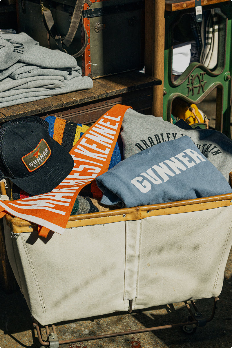A canvas cart filled with folded sweatshirts, an orange and white LIVELINESS scarf, and a black GUNNER cap—essentials for a day at the dog kennel—rests among more clothes and a vintage suitcase. Sunlight casts shadows across the scene.