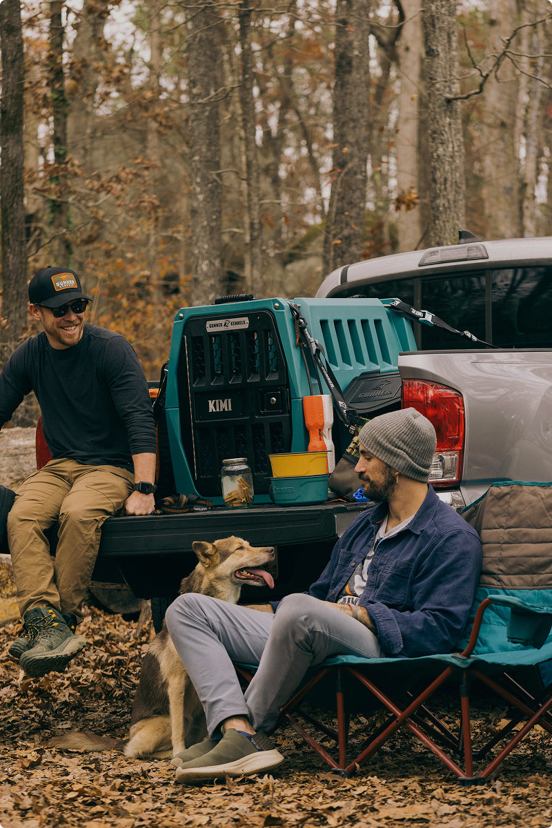 Two men relax at a campsite beside a pickup truck, with one sitting on the bed and the other in a camping chair. A hunting dog sits nearby, surrounded by camping gear on the truck. Trees and fallen leaves fill the background.