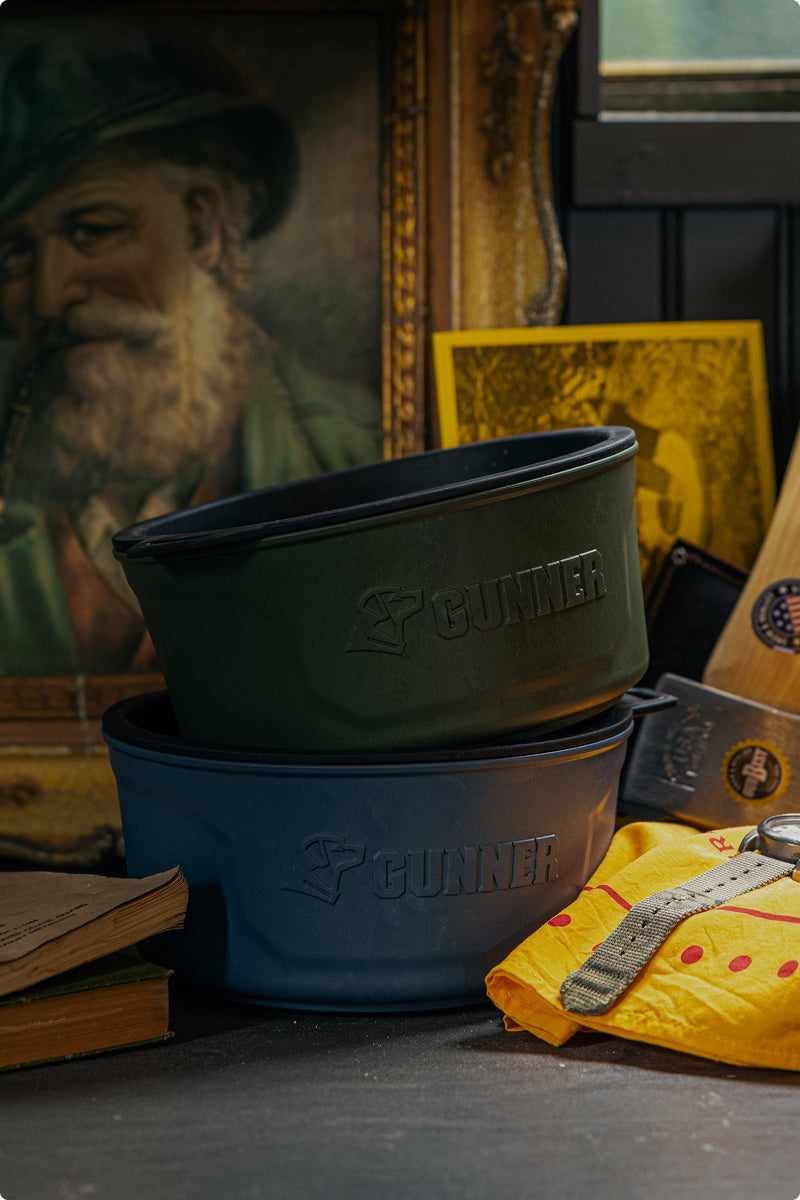 Two stacked GUNNER-branded bowls, one green and one blue, sit on a table with books, a wristwatch, a yellow cloth, and vintage photos—including one of a hunting dog—beside the portrait of a bearded man in the background.