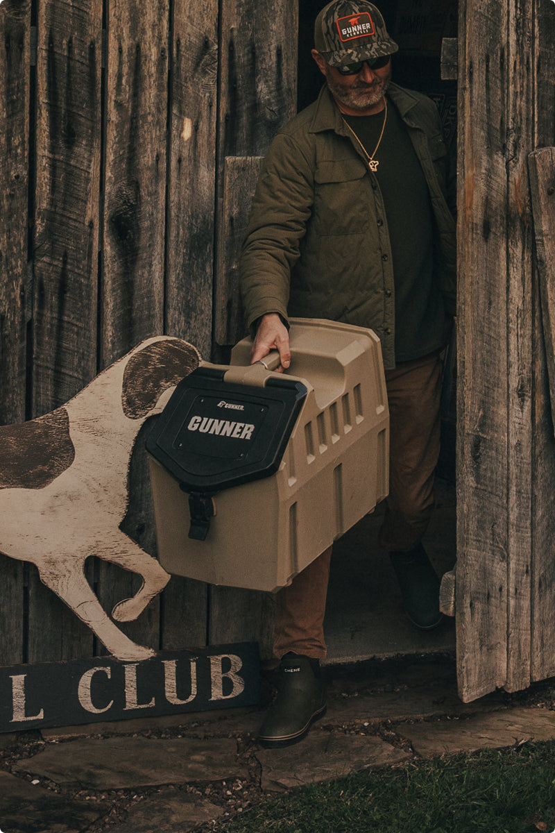 A man in a green jacket and cap carries a tan GUNNER dog crate—essential outdoor gear for any hunting dog—out of a rustic wooden building. A painted dog sign and part of a “CLUB” sign are visible by the door.