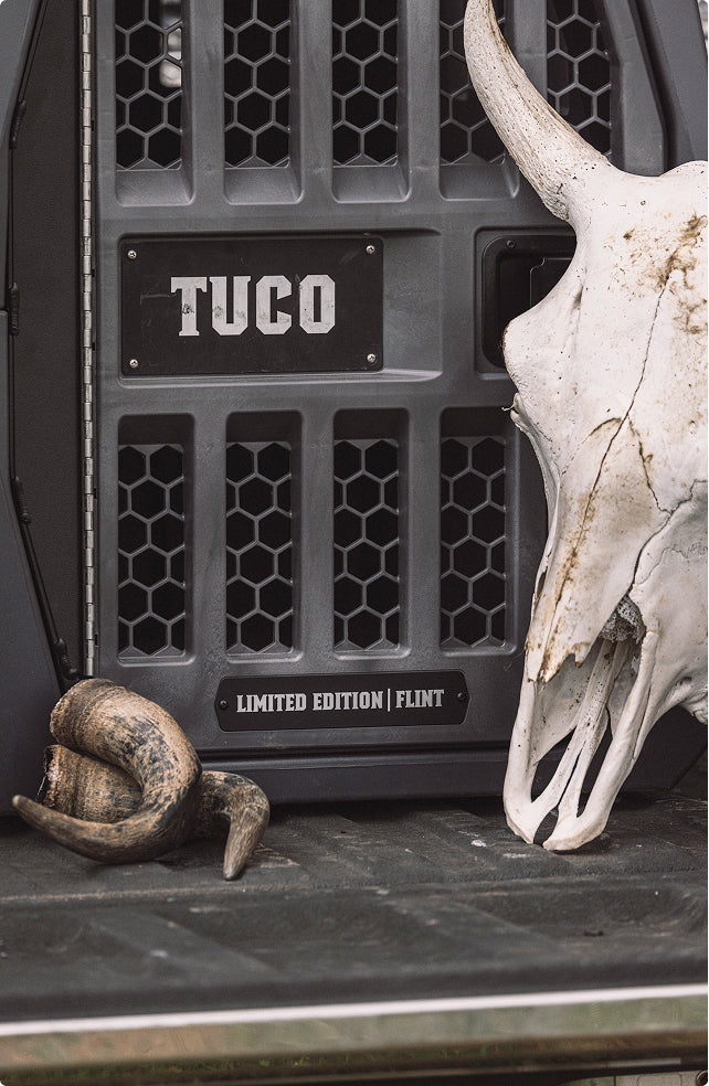 A metal crate labeled TUCO and LIMITED EDITION | FLINT, designed as rugged outdoor gear, is next to a large, weathered animal skull and a curved horn resting on a flat surface—evoking the spirit of an adventurous hunting dog’s kennel.