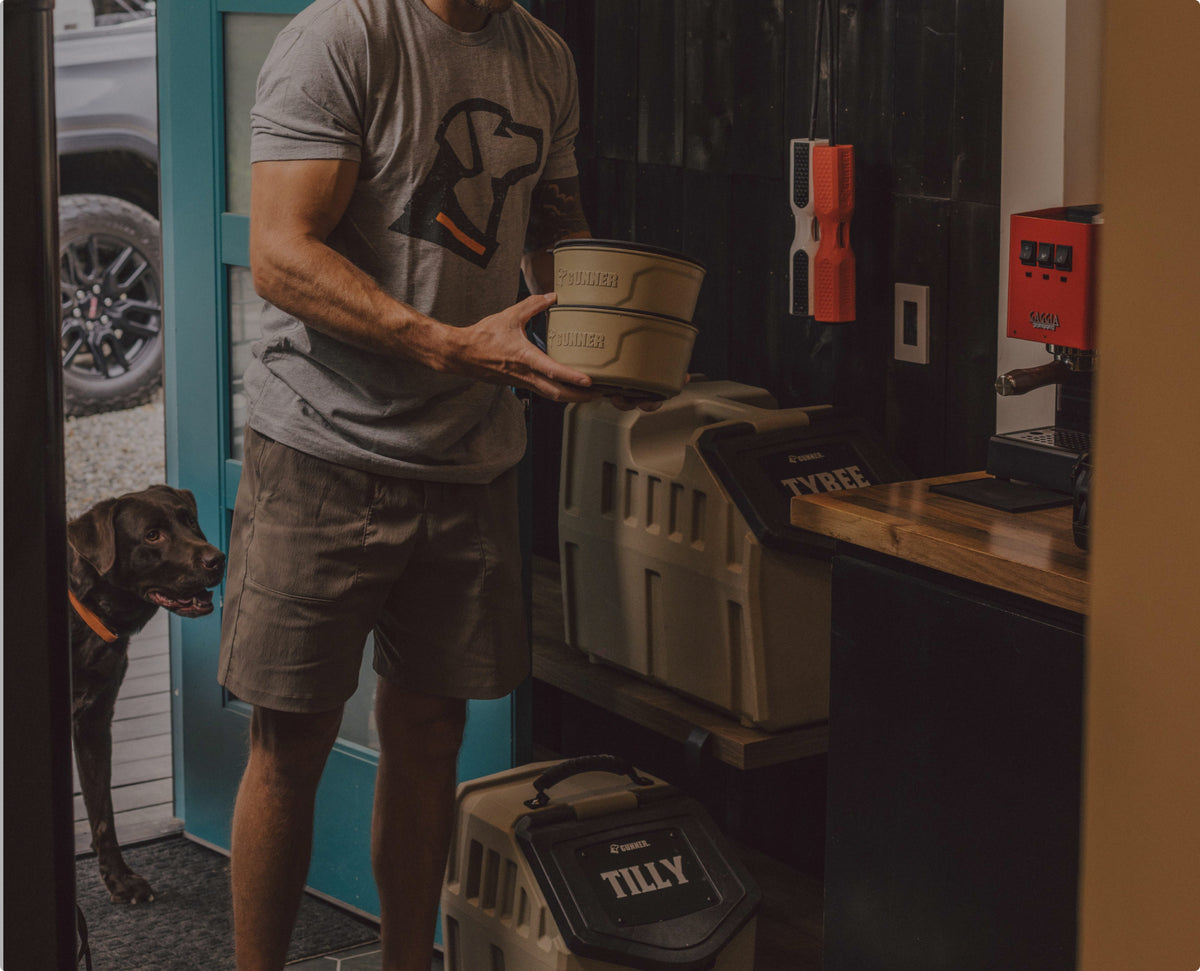 A man in a gray dog-themed t-shirt carries dog bowls labeled CHALLENGE near dog kennels marked TILLY and TYREE, while a brown hunting dog with an orange collar stands by an open door.