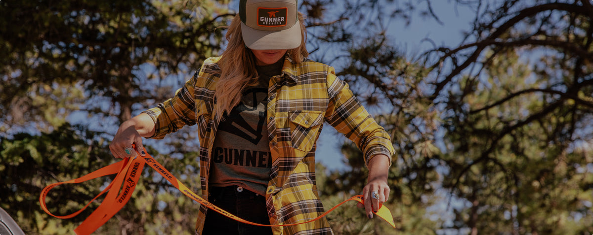 A person wearing a yellow plaid shirt, Gunner t-shirt, and Gunner cap holds an orange leash beside their hunting dog outdoors among trees on a sunny day.