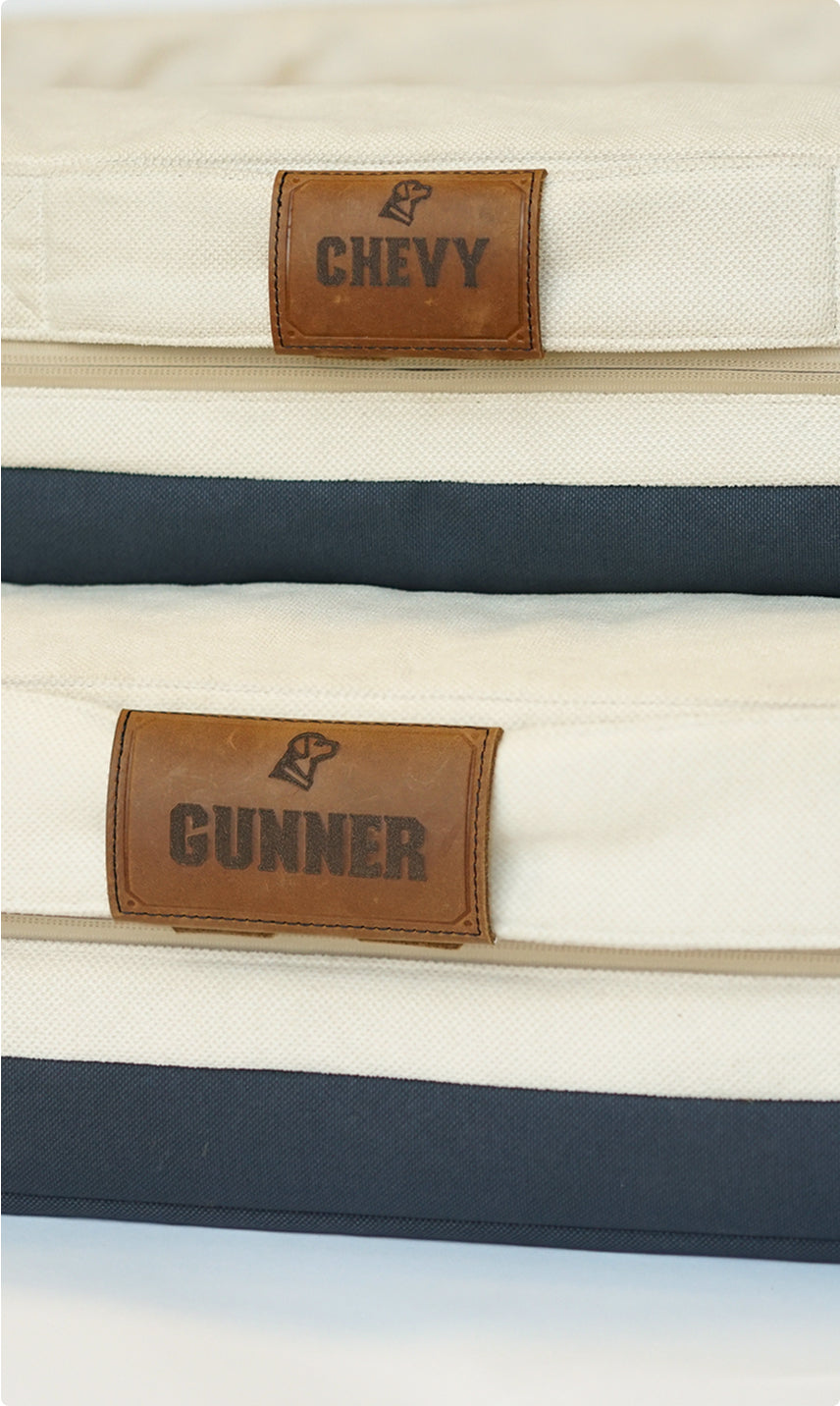 Two stacked canvas cushions with leather name tags—CHEVY on top and GUNNER below—each featuring a small dog paw print icon. Perfect for a cozy dog kennel or for your hardworking, loyal working dog.