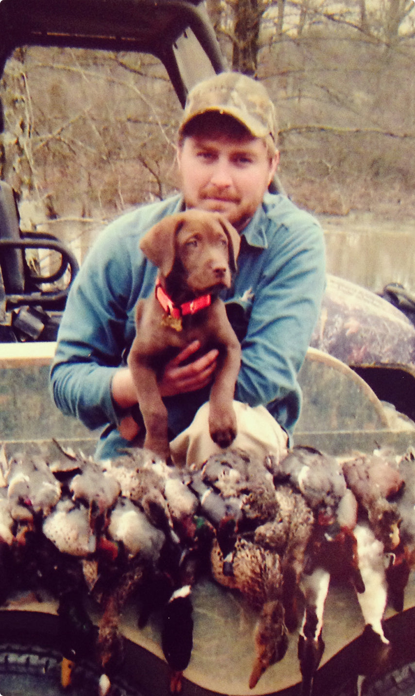 A man wearing a cap holds a brown hunting dog puppy in his lap while sitting on a vehicle. In front of them is a row of several hunted ducks. Leafless trees and water are visible in the background.