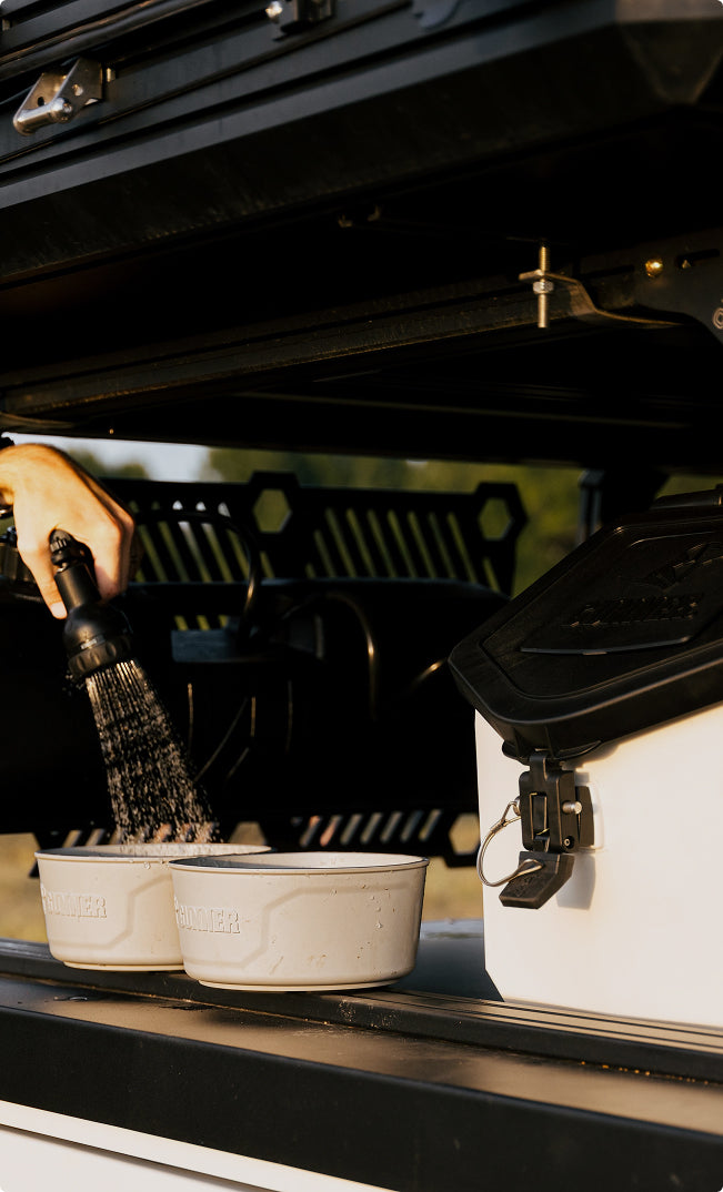 A close-up of a person spraying water into two white bowls on the back of a vehicle, next to a dog crate with a black lid and latch. The scene appears to be an outdoor or camping setup.