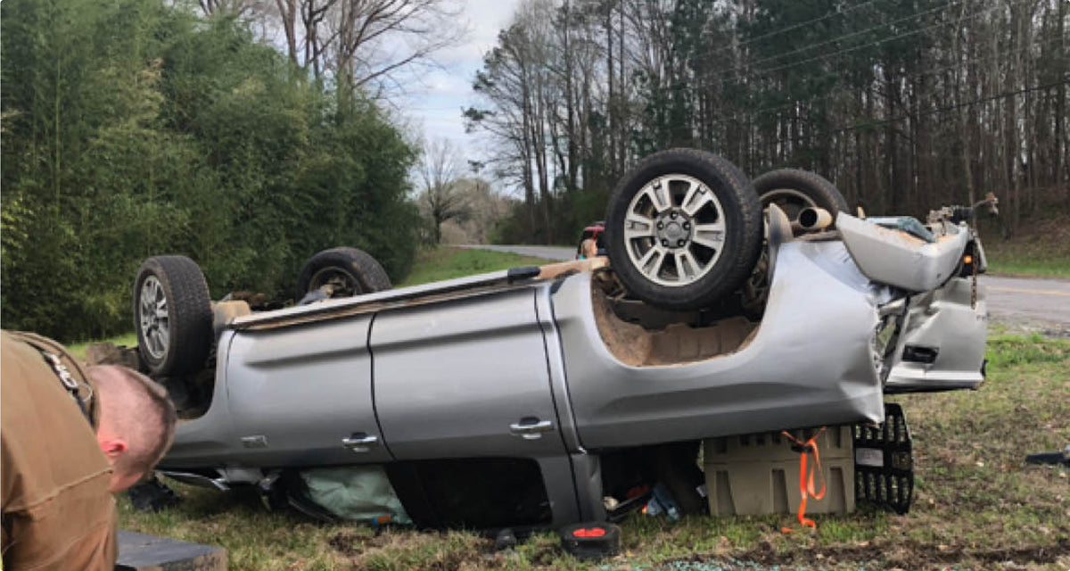 A silver SUV is flipped upside down off the side of a rural road, its wheels in the air and visible damage to the rear. A person, possibly a first responder, kneels nearby beside an outdoor gear bag. Trees and grass surround the scene.