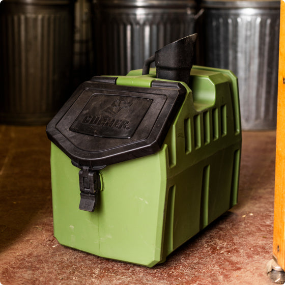 A green plastic container with a black hinged lid and spout sits on a brown floor indoors, near metal trash cans—perfect for use in a kennel or alongside your working dogs crate.