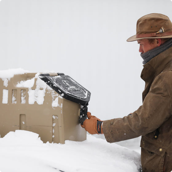 A person in outdoor gear—brown hat, coat, scarf, and gloves—opens a latch on a beige container covered in snow, with a working dog nearby against a white snowy background.