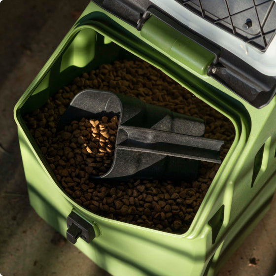A green plastic container filled with brown pet food kibble, perfect for your hunting dog, with a black scoop resting on top of the food inside the open container.