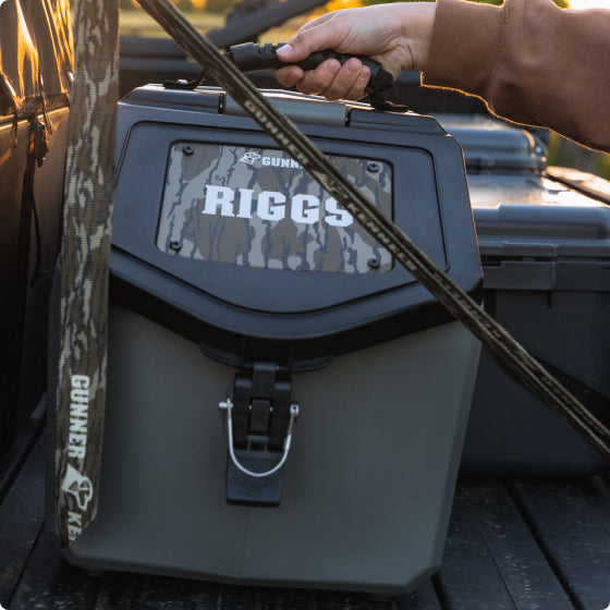 A person’s hand grips the handle of a rugged, camo-patterned dog crate labeled “RIGGS” with “GUNNER” branding, sitting in the back of a vehicle with sunlight in the background—essential outdoor gear for any adventure.