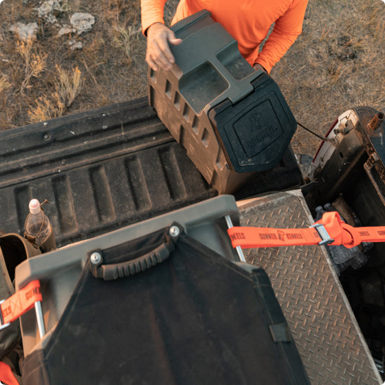 A person in an orange shirt loads a large black kennel onto the back of a pickup truck with a textured metal bed and orange straps, outdoors on dry grass, preparing outdoor gear for their working dog.