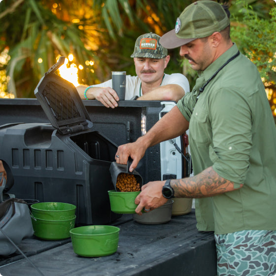 Two men stand at the back of a pickup truck. One scoops kibble into green dog bowls for a hunting dog, while the other watches and holds a mug. Trees and sunlight are visible in the background.