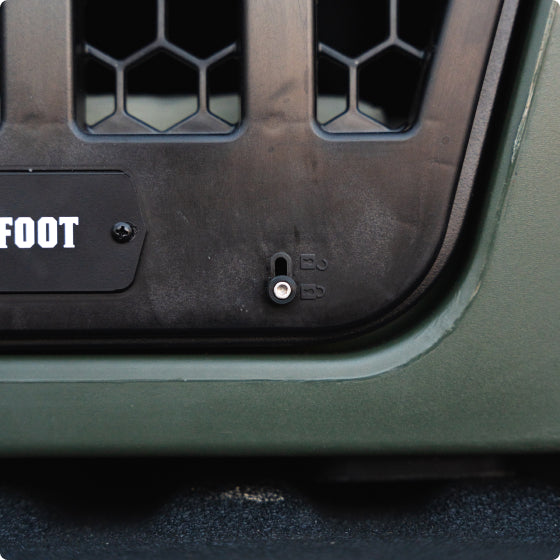 Close-up of a dark green and black dog crate with a vented panel and small circular metal lock. The word FOOT is partially visible on a label to the left. The kennel’s surface has a matte texture.
