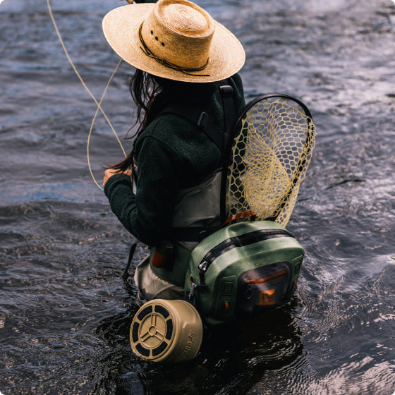 Person wearing a straw hat and outdoor gear stands knee-deep in water, holding a fishing rod. A fishing net and green backpack are strapped to their back as the water flows gently around them.