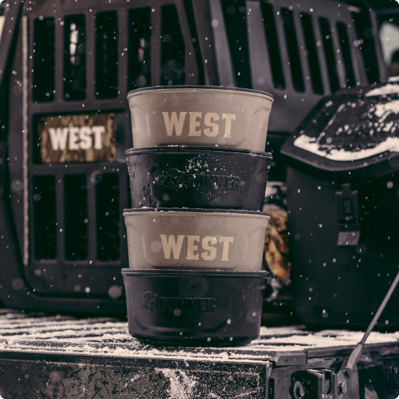 Three stacked dog bowls labeled WEST sit on a snowy truck bed. A large working dog kennel with the word WEST is in the background, and snow is falling around the scene.
