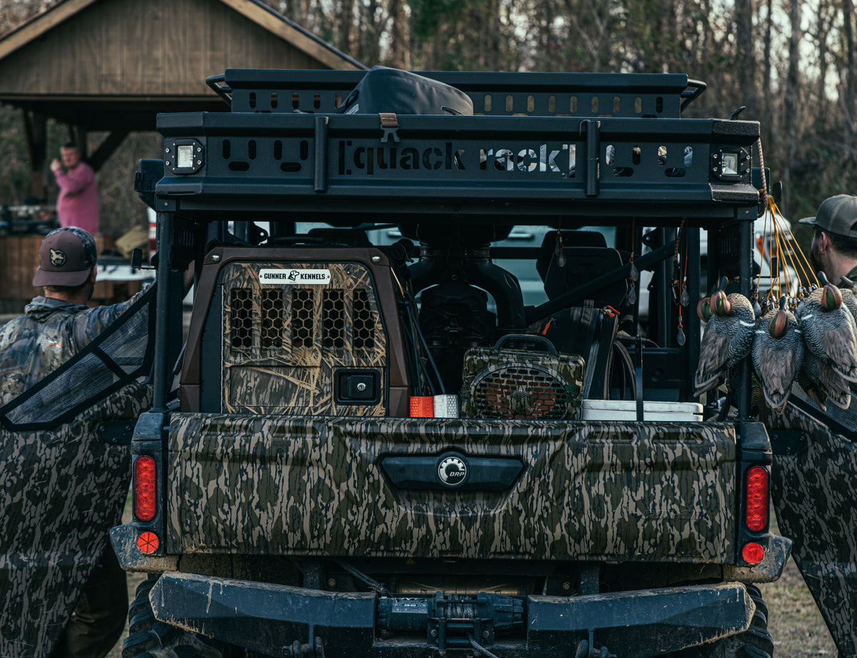 A GUNNER G1™ KENNEL in Susquehanna/Mossy Oak Shadow Grass sits in a camouflaged off-road vehicle packed with hunting equipment. Two camo-clad duck and goose hunters stand nearby; another person waits by a wooden shelter in the background.