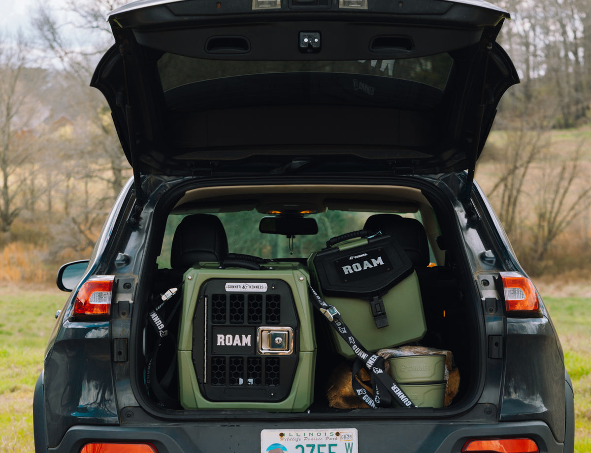 The open trunk of an SUV reveals GUNNER gear, including the G1™ KENNEL - GUNNER GREEN, arranged neatly. The car is parked outdoors with trees and grass in the background.