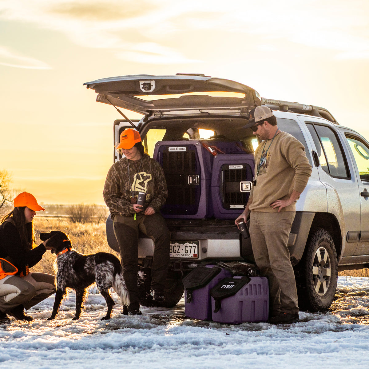 Three hunters in orange hats stand by an SUV at sunset, chatting and petting a black and white dog after a run. The snow-covered trunk is filled with gear, including the GUNNER G1™ KENNEL - MOSSY OAK BOTTOMLAND DUSK.
