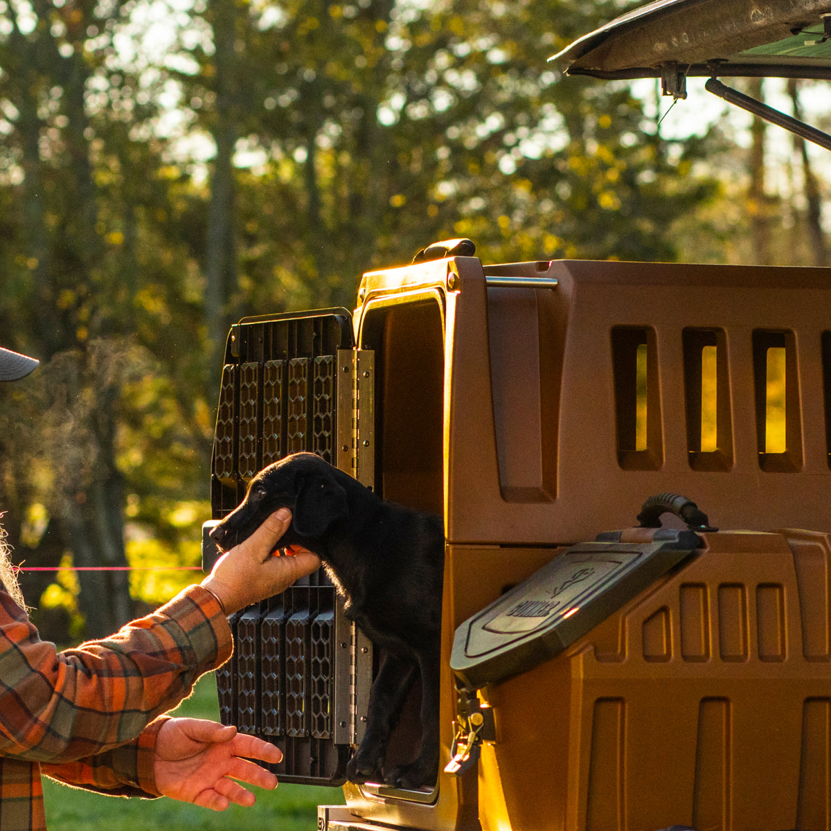 A person in a plaid shirt pets a black dog as it steps out of an open brown GUNNER G1™ KENNEL - SUSQUEHANNA, attached to a vehicle amid trees and sunlight with hints of Flyway colors in the background.