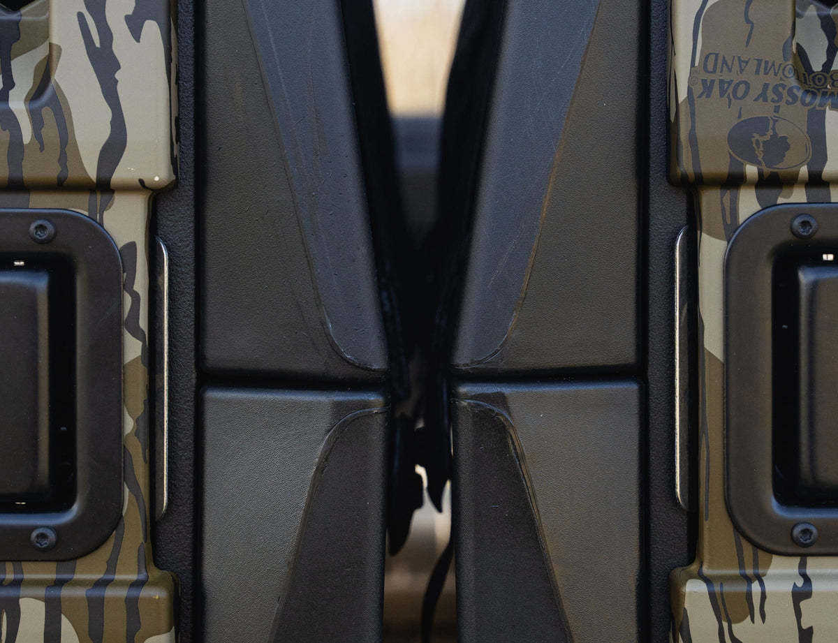 Close-up of two camouflage-patterned vehicle doors nearly touching, with black handles and hinges, their rugged surfaces forming a narrow gap—evoking the secure build of the GUNNER G1™ KENNEL - MOSSY OAK BOTTOMLAND BLACK, designed to protect dogs’ lives.