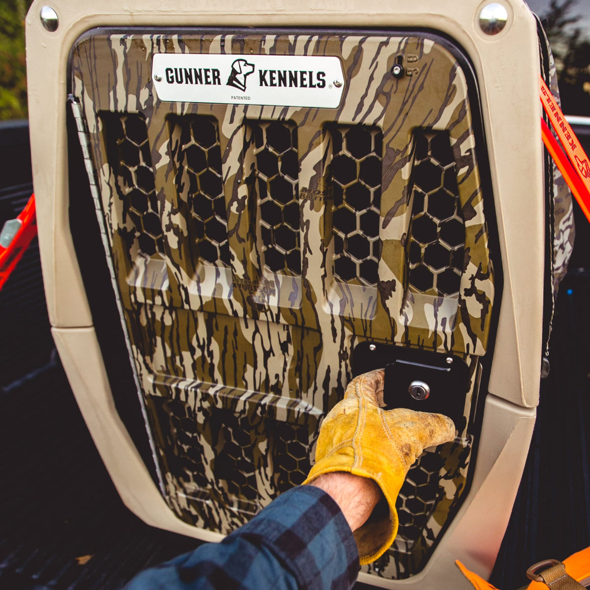Wearing a yellow work glove, someone secures a GUNNER G1™ KENNEL in Mossy Oak Bottomland Tan in the back of a vehicle using red straps.