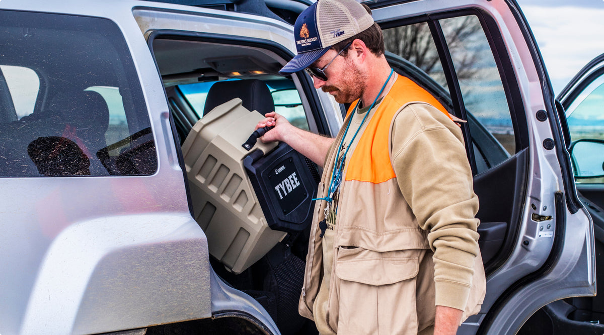A man wearing sunglasses and a cap loads a large beige kennel labeled TYDEE into the backseat of a silver SUV, ready to transport his working dog and outdoor gear.