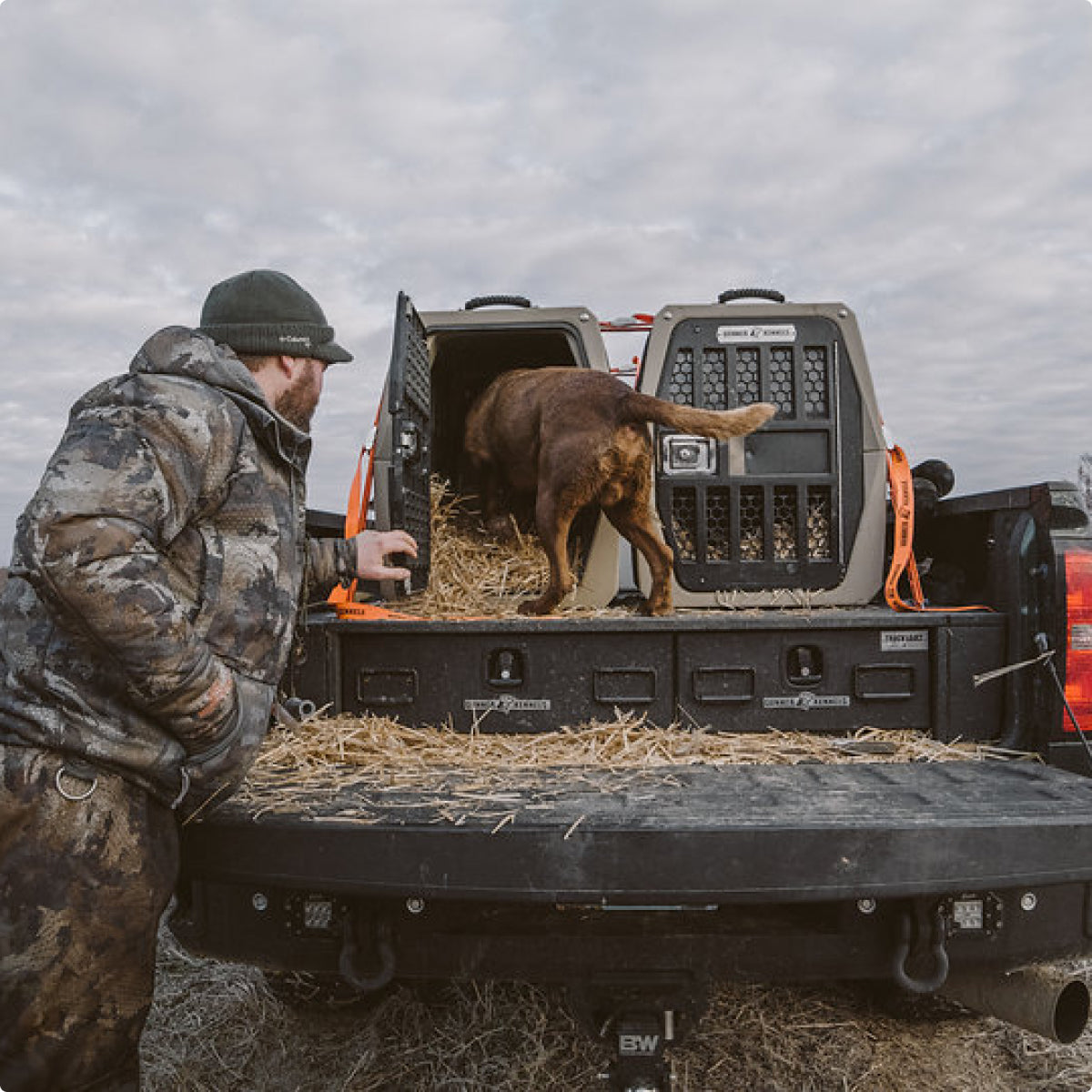 A person in camouflage outdoor gear stands by a pickup truck as a brown dog enters a dog crate on the straw-lined truck bed under a cloudy sky.