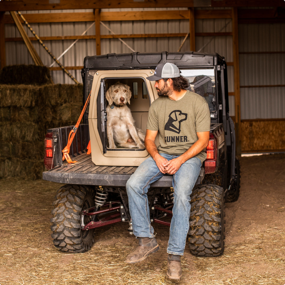 A man in a cap and GUNNER t-shirt sits on the tailgate of an off-road vehicle, looking at a dog resting inside a dog crate in the back, surrounded by hay bales and outdoor gear inside a barn.