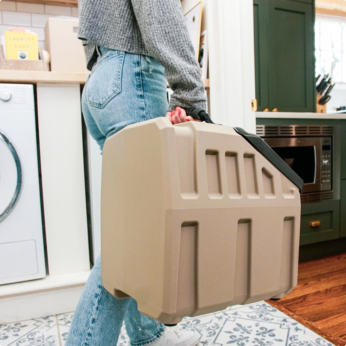 A person in jeans and a gray sweater carries a large, tan, rectangular plastic kennel with a handle in a kitchen. A washing machine, green cabinets, and a microwave are visible in the background.