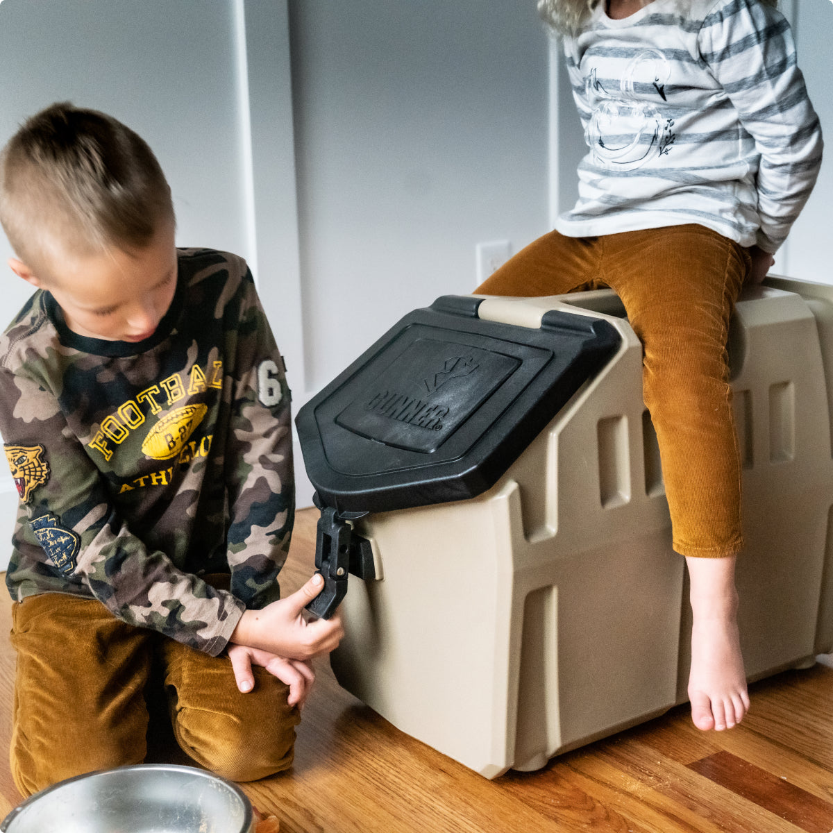 Two children in casual clothes interact with a large beige plastic dog kennel; one sits on top while the other fastens the latch, with a metal bowl resting on the wooden floor nearby.