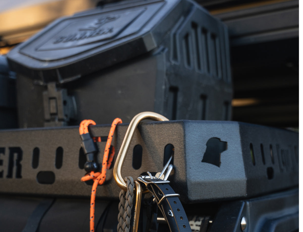 Close-up of outdoor gear including a black utility box, an orange and black rope, a gold carabiner, and a braided black strap—essentials for any hunting dog owner—hanging from a perforated metal panel.