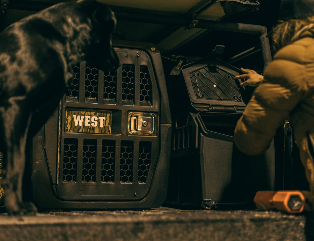 A person in a yellow jacket opens a large black dog crate attached to a vehicle, with a black dog nearby. Next to the crate is the GUNNER FOOD CRATE - Black by GUNNER, keeping the dogs food fresh and pest-free outdoors at night.