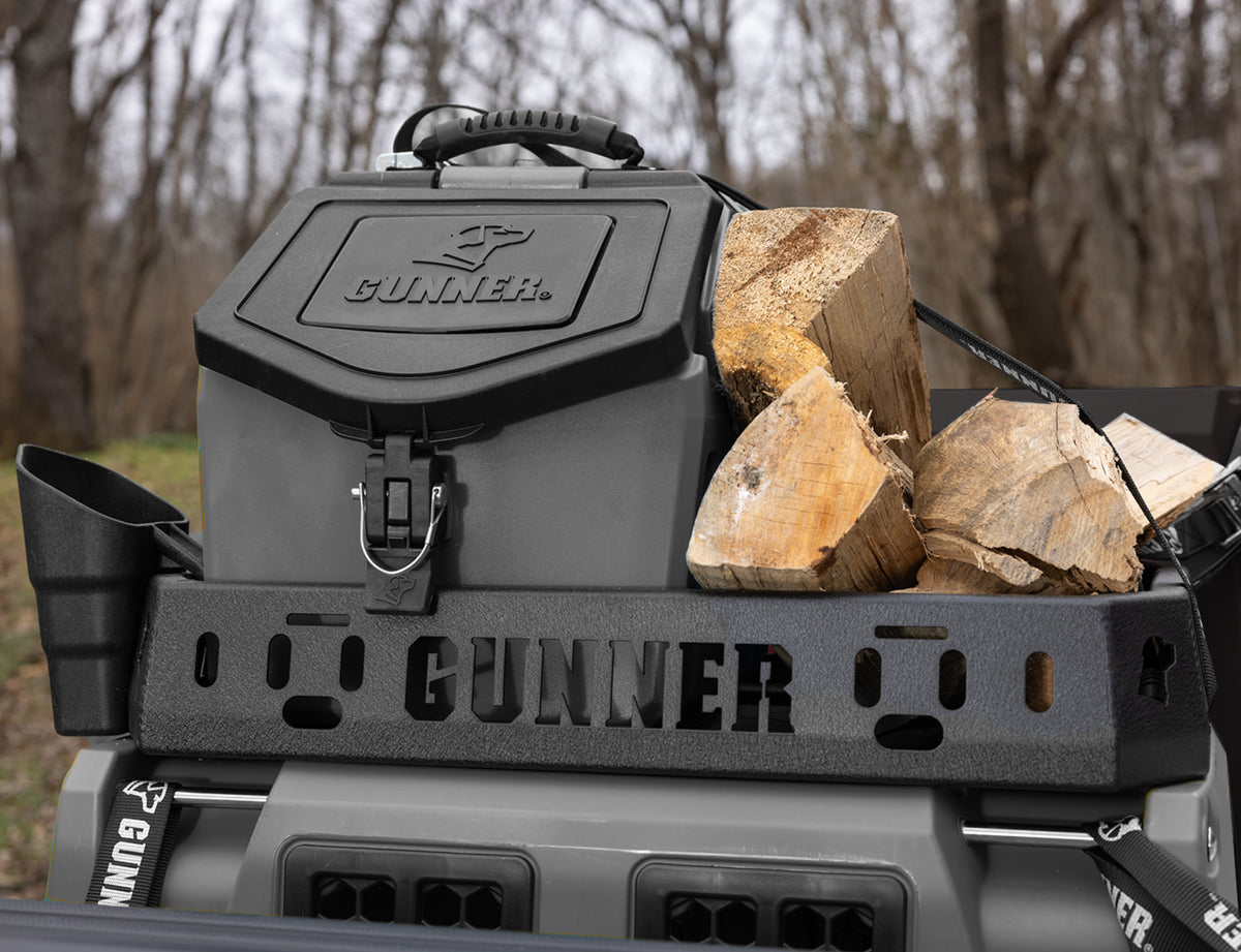 A black GUNNER® storage box, perfect for hunting dog gear, and several pieces of chopped firewood are secured in a truck bed rack marked “GUNNER,” with trees and grass visible in the background.