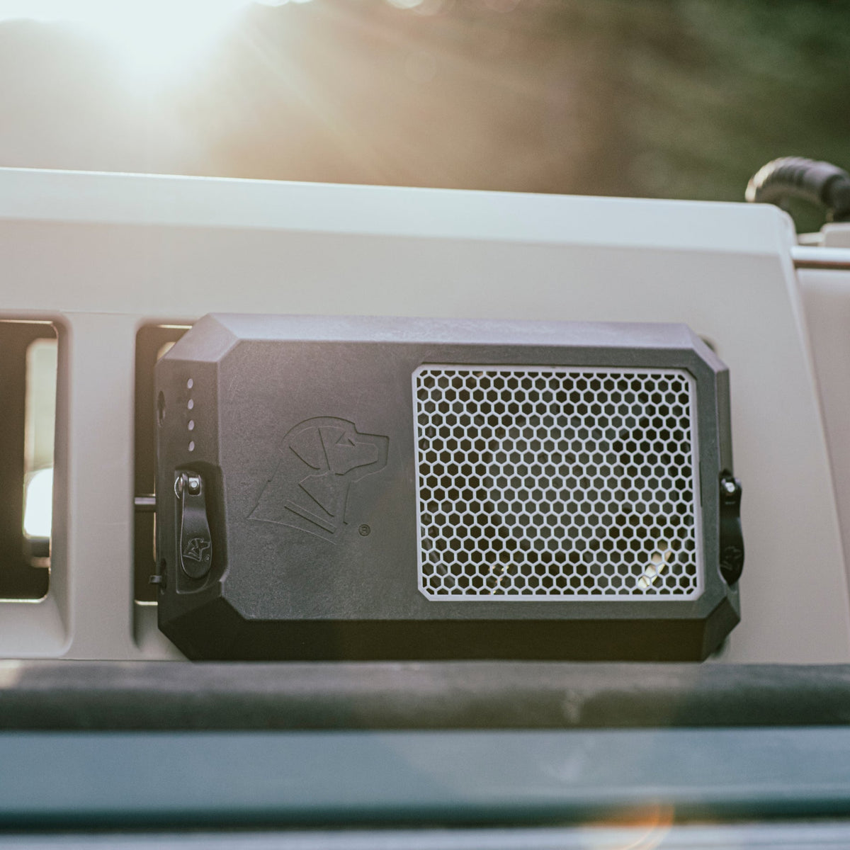 A close-up of a black, vented dog crate door with honeycomb mesh and an embossed dog head, attached to a vehicle in sunlight. Compatible with GUNNER accessories like the GUNNER FAN KIT for weatherproof cooling.