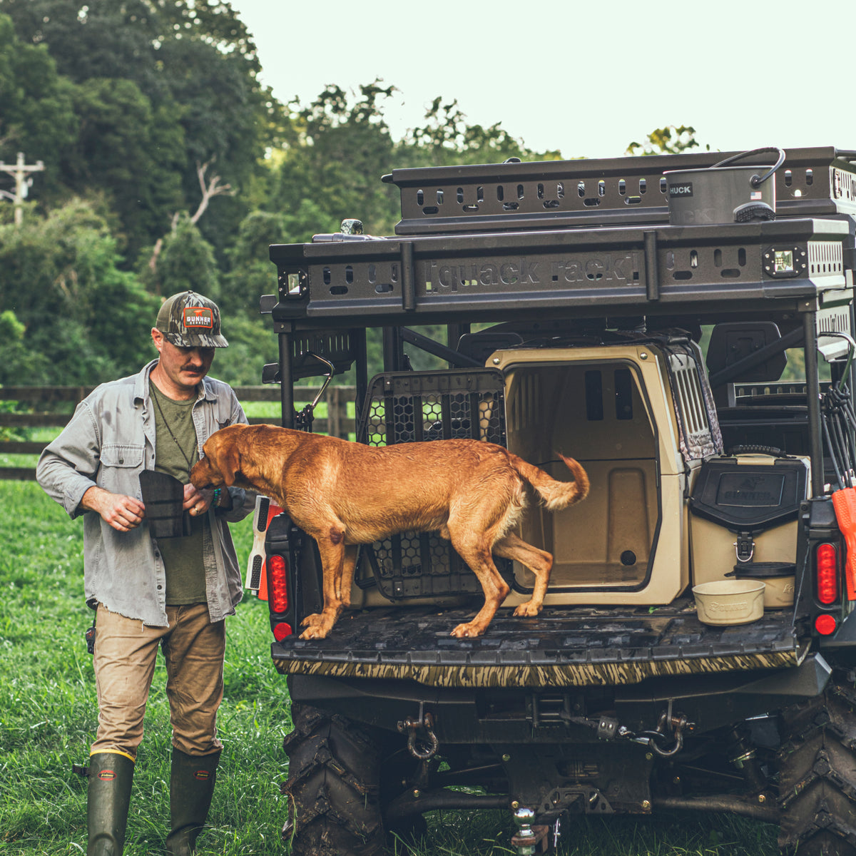 A man in outdoor gear stands by an off-road vehicle with a brown dog on the tailgate next to a GUNNER FOOD CRATE - TAN by GUNNER, in a grassy field with trees behind them.