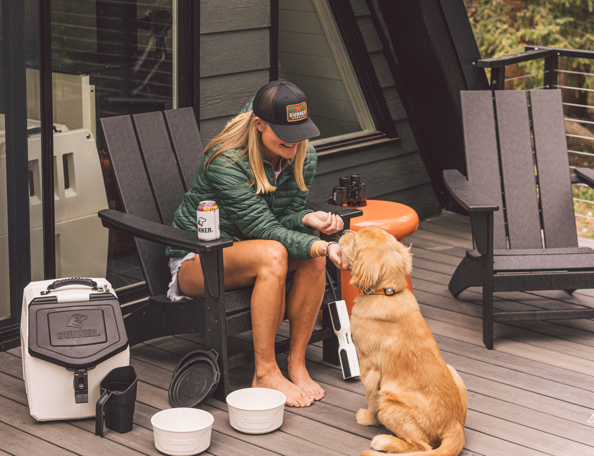 A woman in a green jacket and the GUNNER BLACK MALLARD TRUCKER hat sits on a porch chair feeding treats to a golden retriever. Pet bowls, a cooler, and a drink are nearby. The setting is outdoors beside a modern cabin.