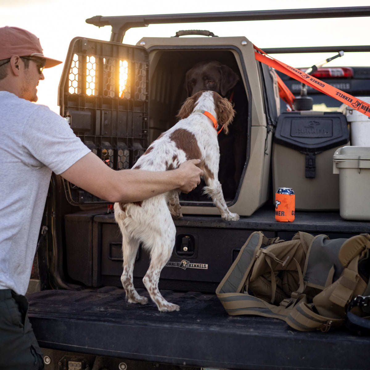 A man helps a brown and white dog into a truck bed kennel where another brown dog waits. On the tailgate are camping gear, a cooler, and the GUNNER FOOD CRATE - TAN by GUNNER as the sun sets behind them.