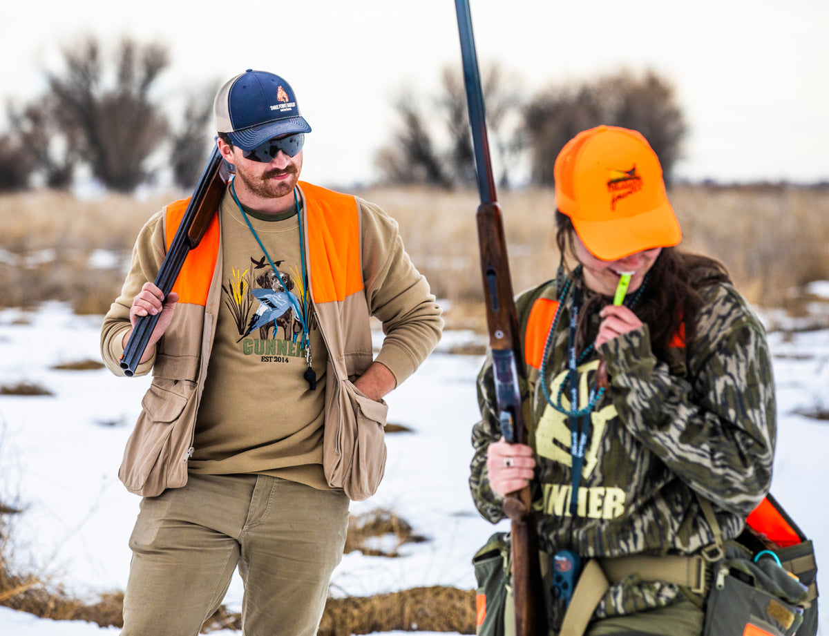 Two hunters in orange vests stand in a snowy field. The man holds a shotgun, while the woman, wearing an orange hat and a GUNNER BOTTOMLAND HOODIE, looks down and blows a whistle. Trees and grass fill the background.