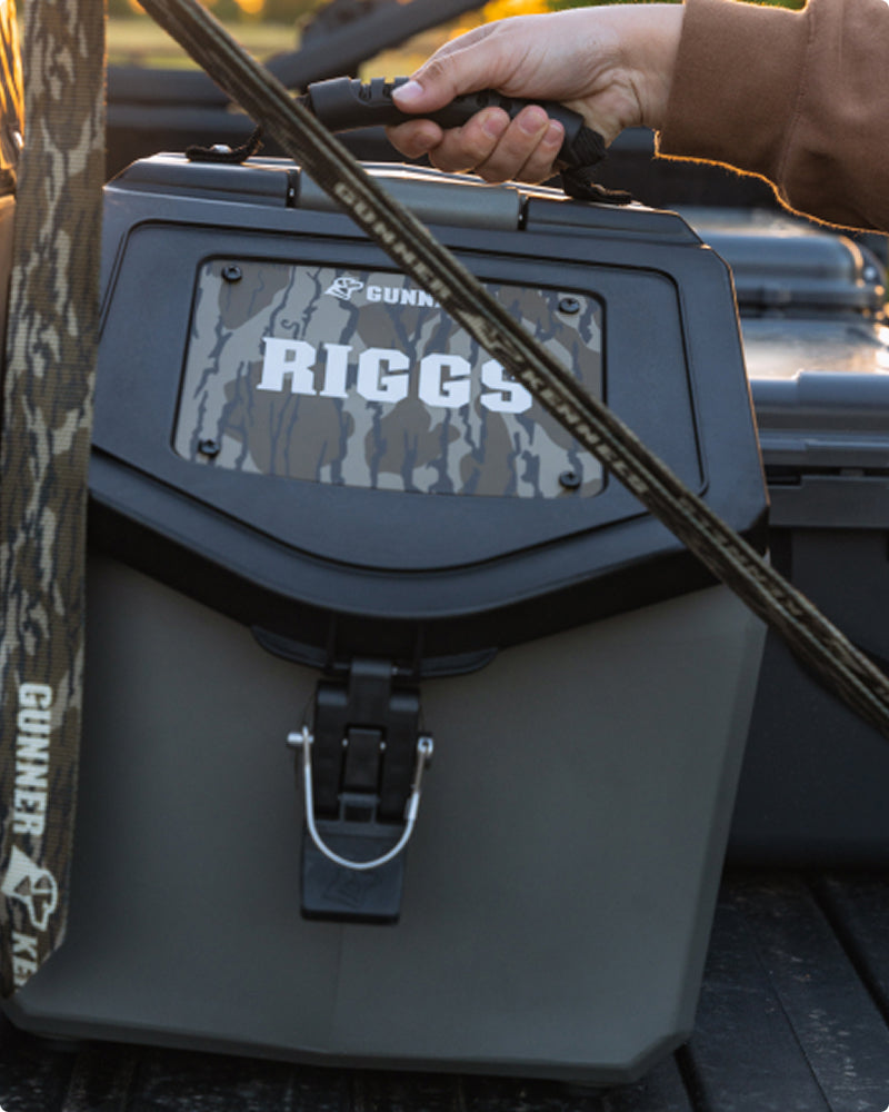 A person’s hand holds the handle of a gray and black cooler with a camouflage panel labeled RIGGS; camouflage straps, perfect for securing to a dog crate or bringing along for hunting dog trips, are draped over the cooler.