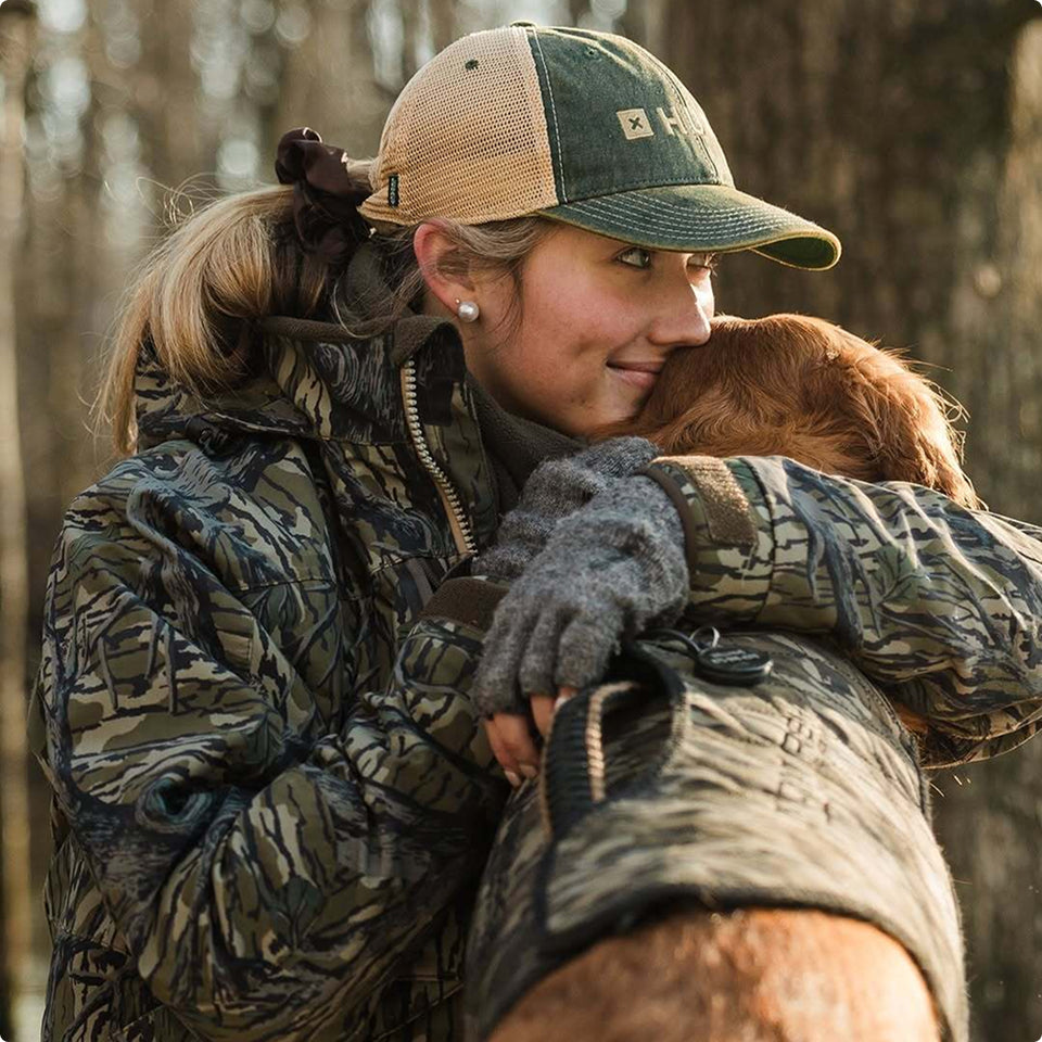 A woman in a camouflage jacket and cap smiles as she hugs her brown hunting dog, both wearing matching coats, while standing outdoors in a sunlit forest.