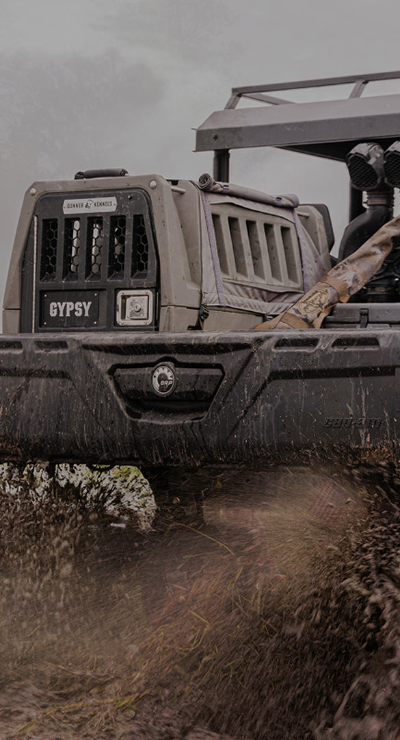 A rugged off-road vehicle labeled GYPSY drives through muddy terrain, splashing mud and water, with outdoor gear secured on top and a dog crate in the back, all set against a cloudy sky.