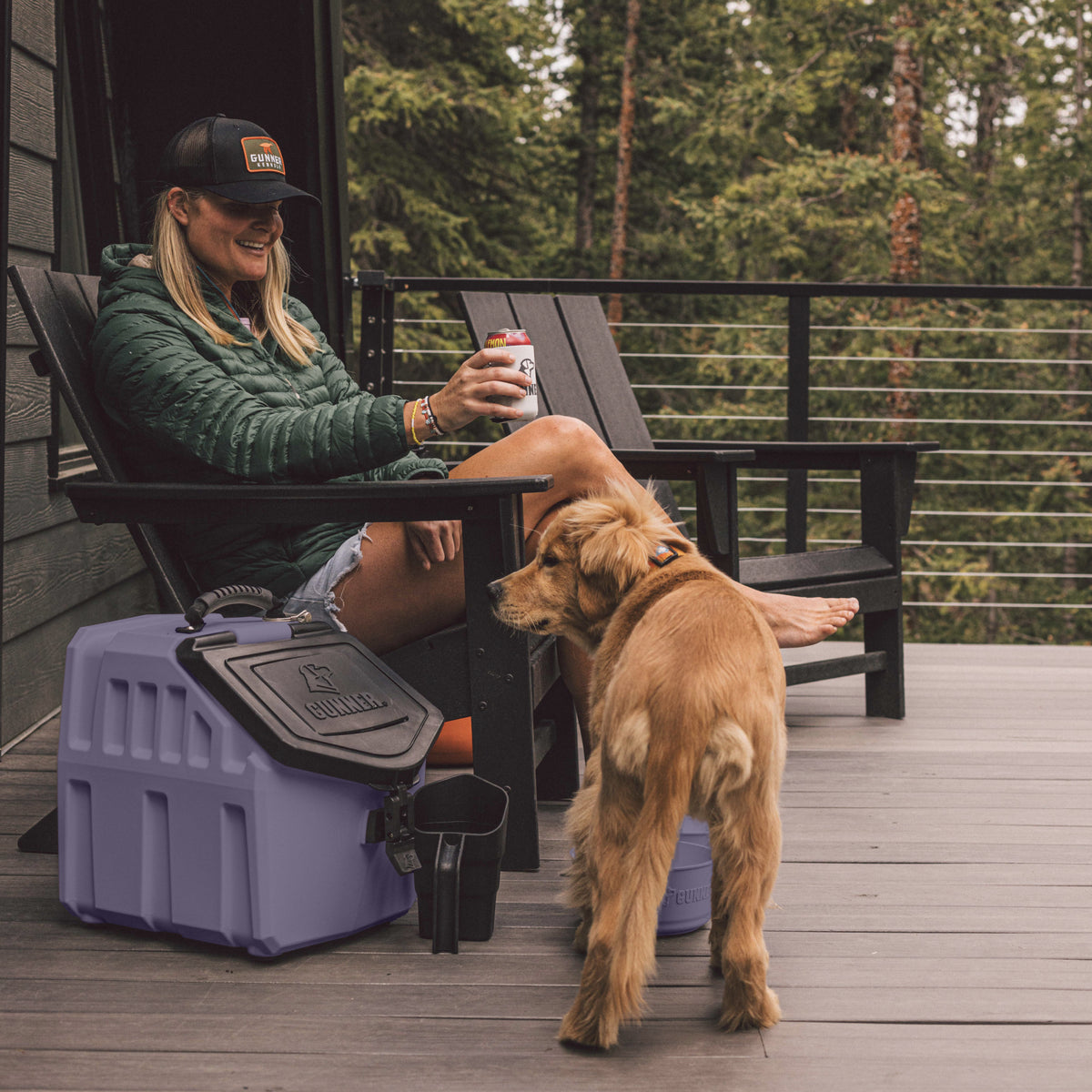 At dusk, a woman in a green jacket and hat relaxes on a deck chair, smiling with a drink as her golden retriever stands nearby beside the GUNNER FOOD CRATE by GUNNER, surrounded by trees and wooden decking.
