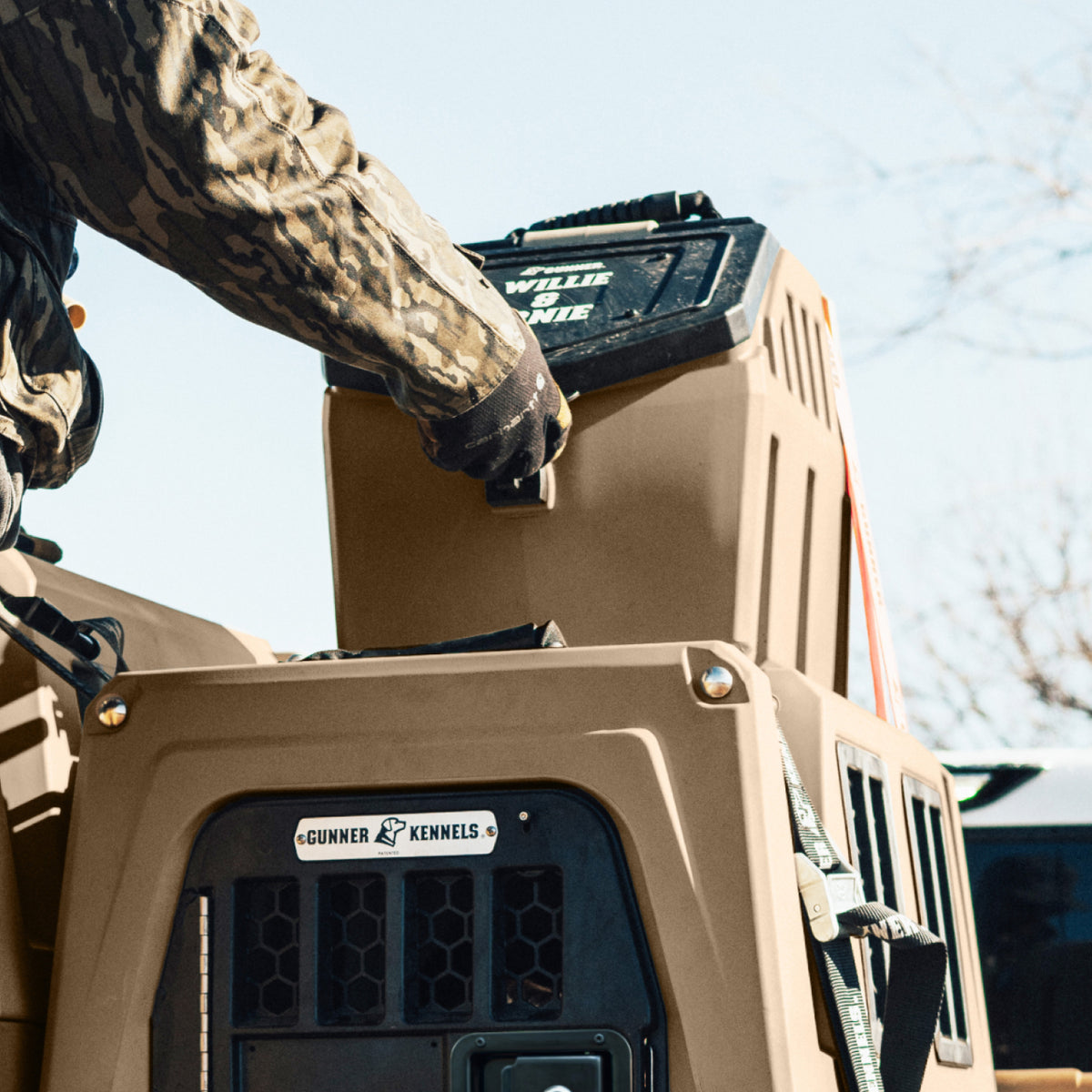 A person in camouflage clothing lifts a GUNNER FOOD CRATE - Cheyenne Bottoms onto a tan GUNNER dog kennel outdoors under a clear sky.