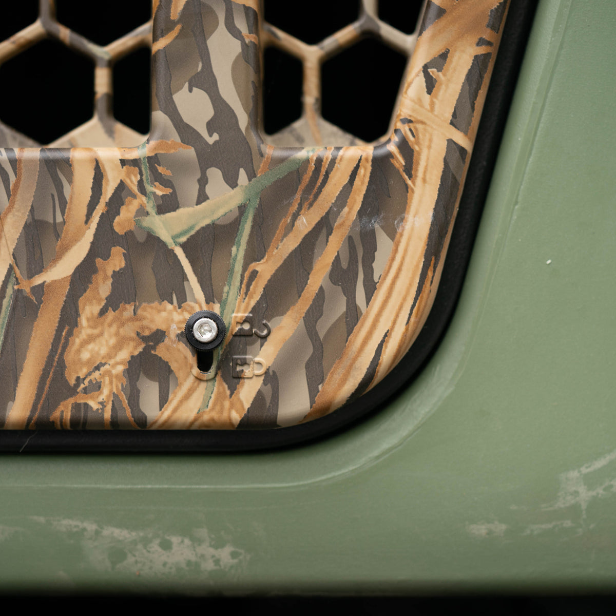 Close-up of a vehicle grille with a camouflage pattern, honeycomb mesh, and a bolt—ideal for transporting a working dog or hunting dog. The edge of the grille is bordered by a solid green exterior, offering durability whether you’re heading out from the kennel or into the field.