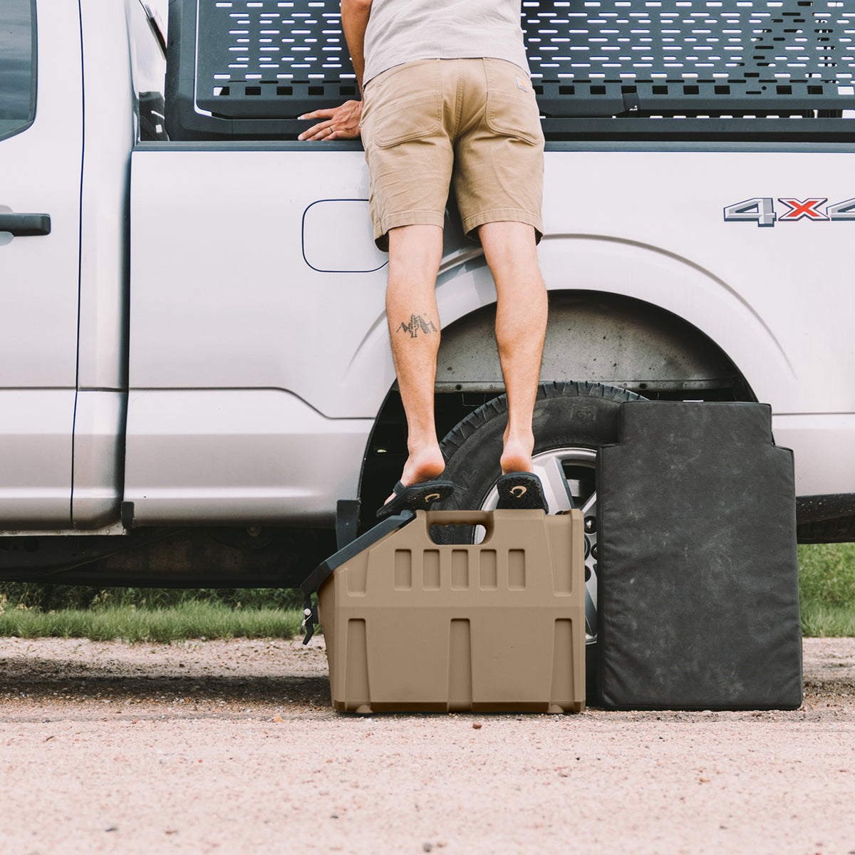 A person in shorts stands on a GUNNER FOOD CRATE - Cheyenne Bottoms by GUNNER to reach into the bed of a silver pickup truck. A black cover and a tire are nearby, with the person’s back and legs visible.